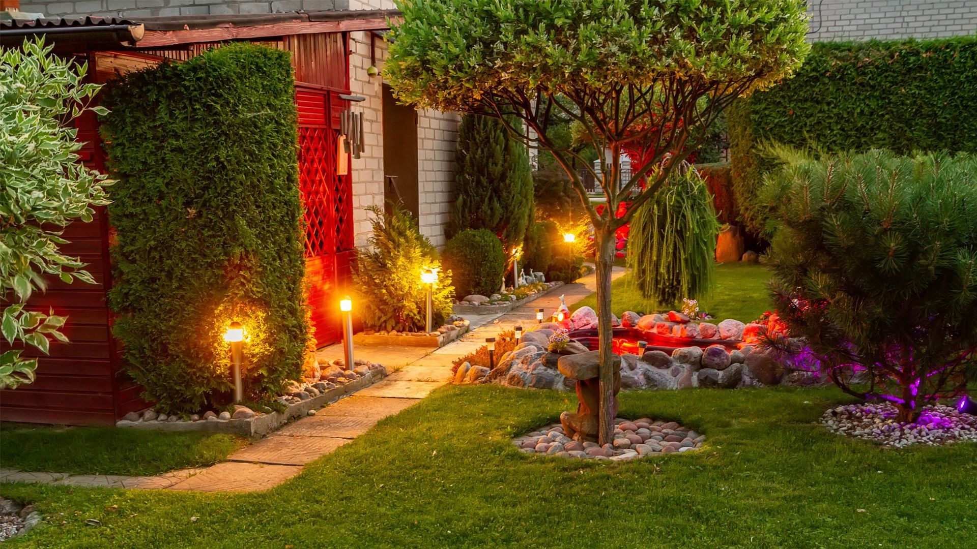 Lit garden path beside a brick house, with flowers, shrubs, and a tree at dusk