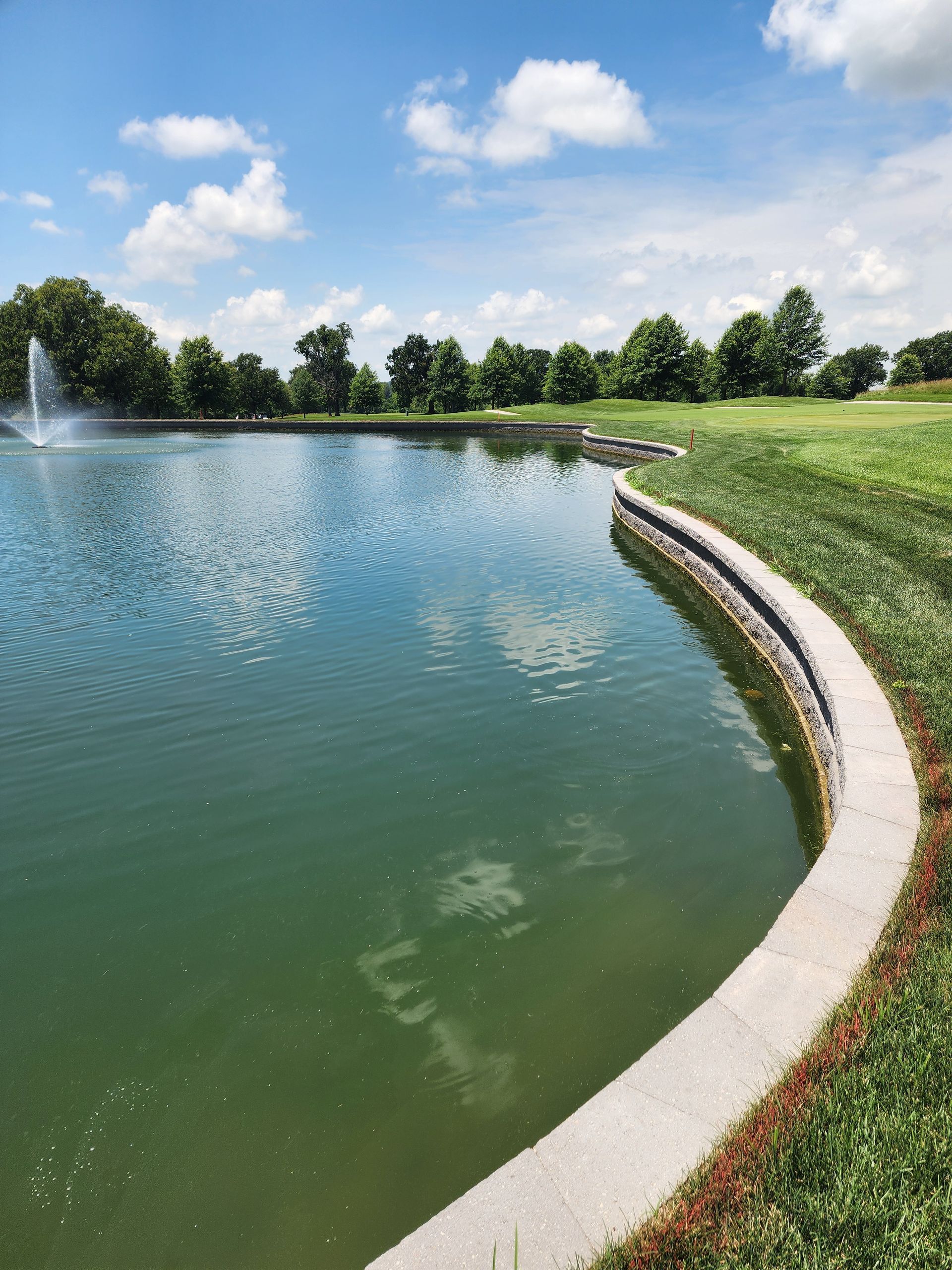 Curved concrete pond edge beside a grassy bank and blue-green water with a fountain under a partly cloudy sky