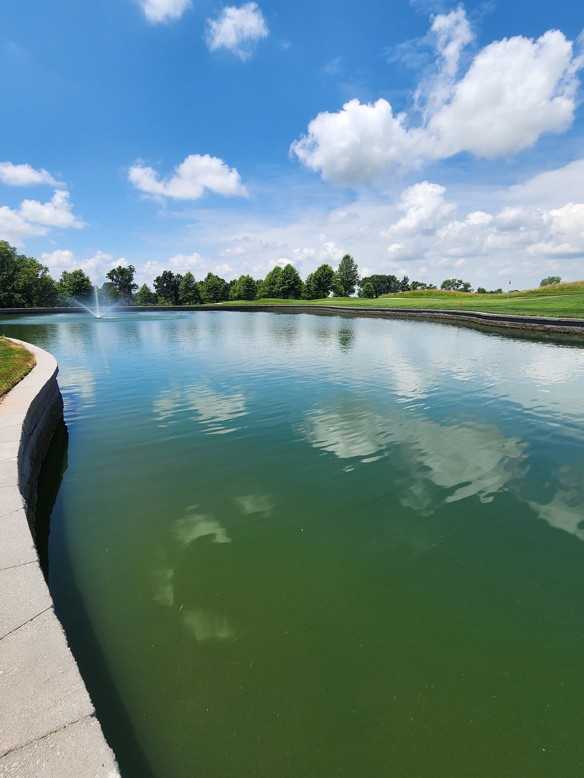 Curved green lake reflecting clouds, with a paved path along the left edge and trees in the distance