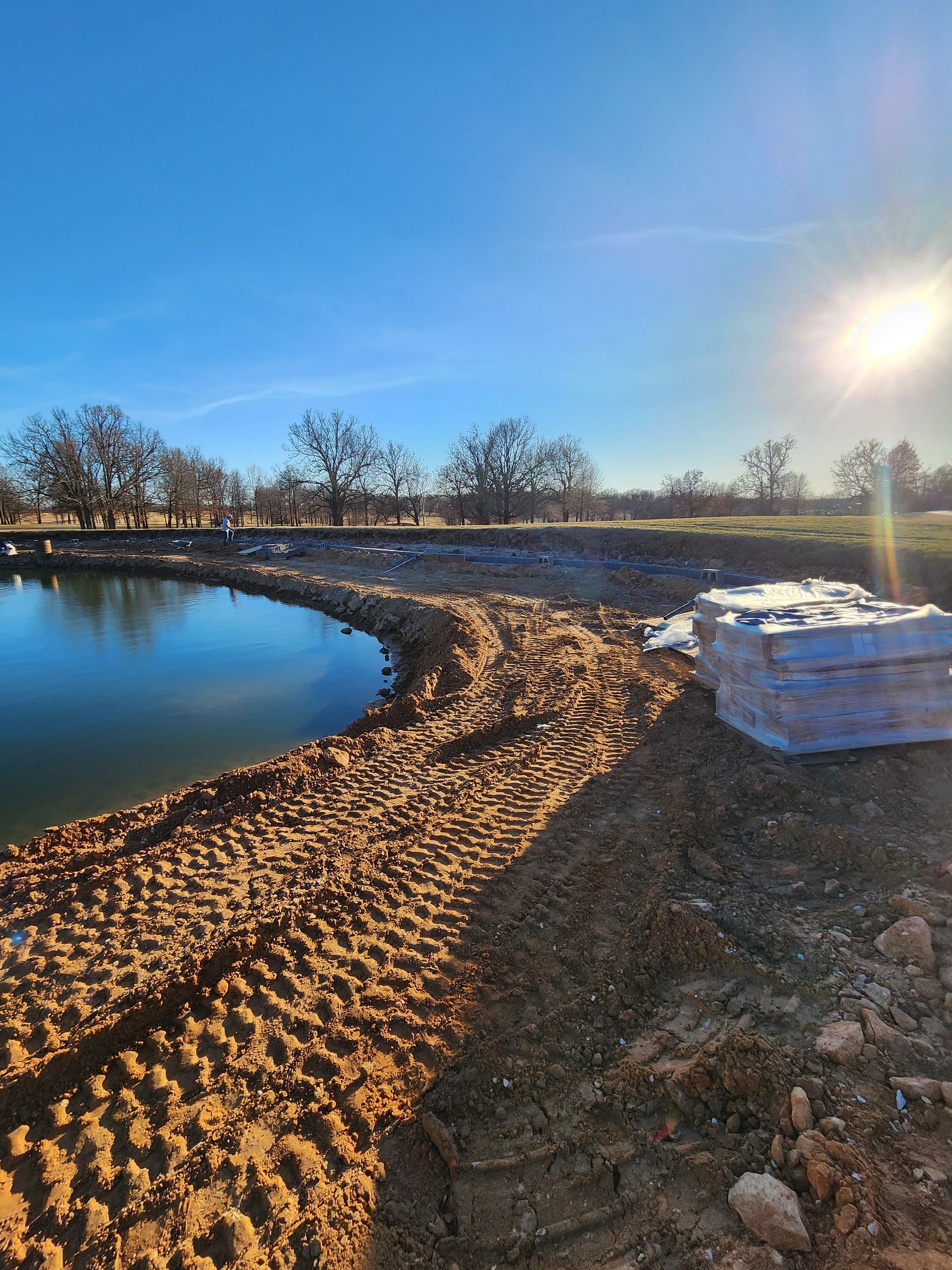 Curving dirt path beside a calm pond under bright sun, with snow along the shore and bare trees in the distance