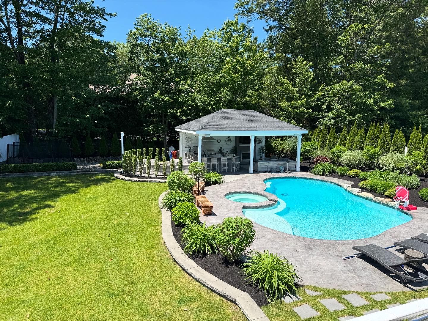 A backyard featuring a turquoise swimming pool with a hot tub, stone patio, and a covered pavilion under sunny skies.