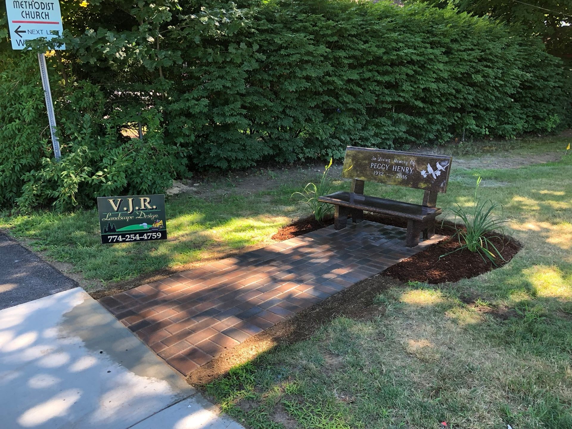 A dark wooden bench sits on a paved path in a grassy area next to a bush, with a small sign nearby.
