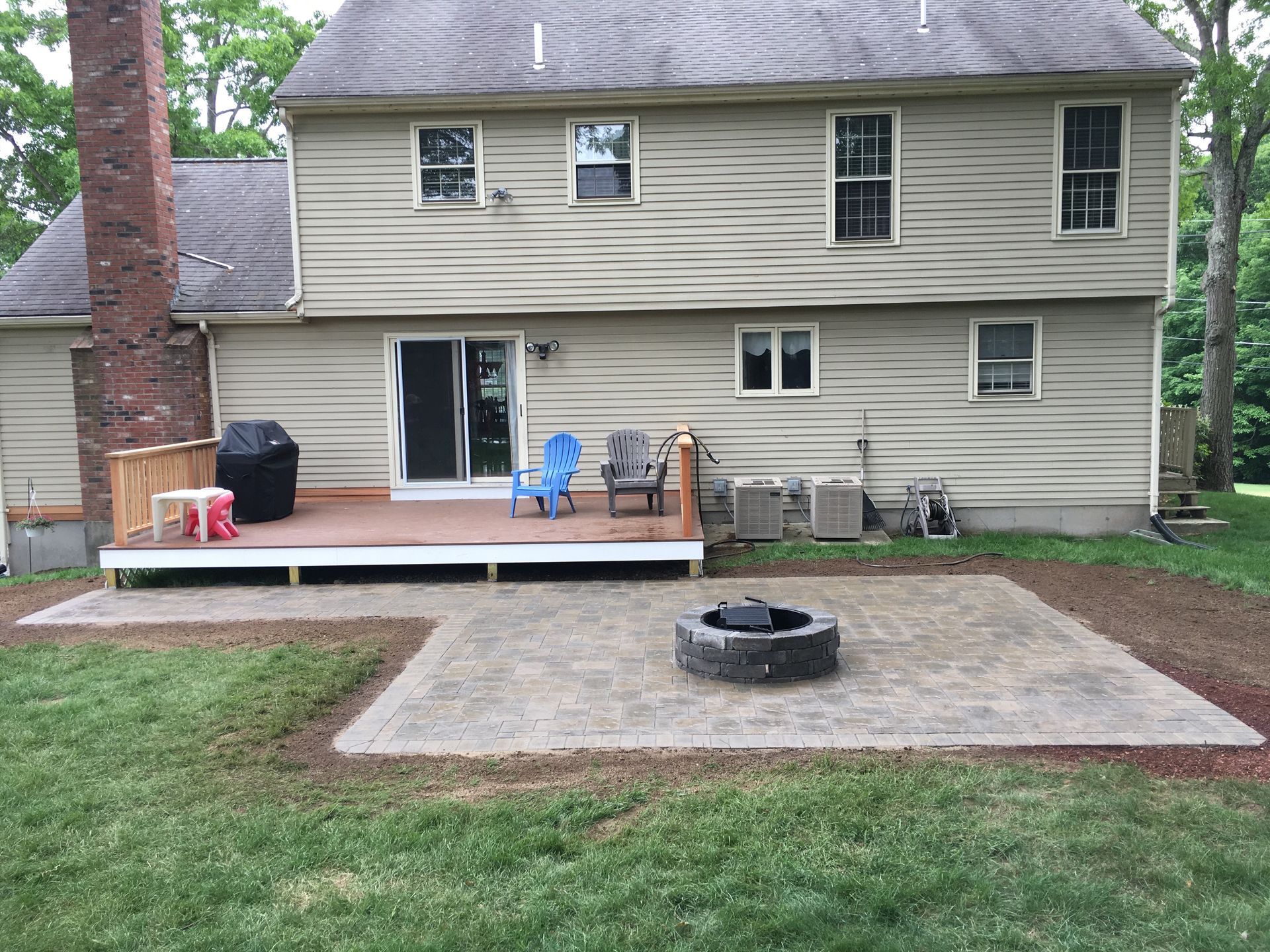 A backyard with a beige two-story house, a wood deck with a grill and chairs, and a paved stone patio with a fire pit.