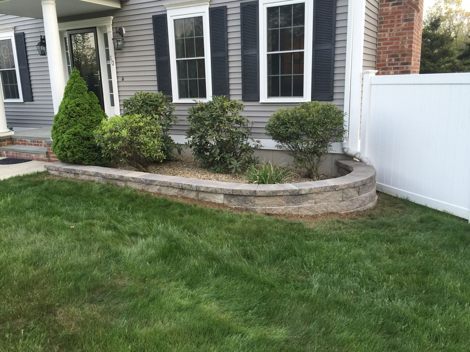 A curved stone retaining wall in front of a gray house with black shutters, landscaping, and a white privacy fence.