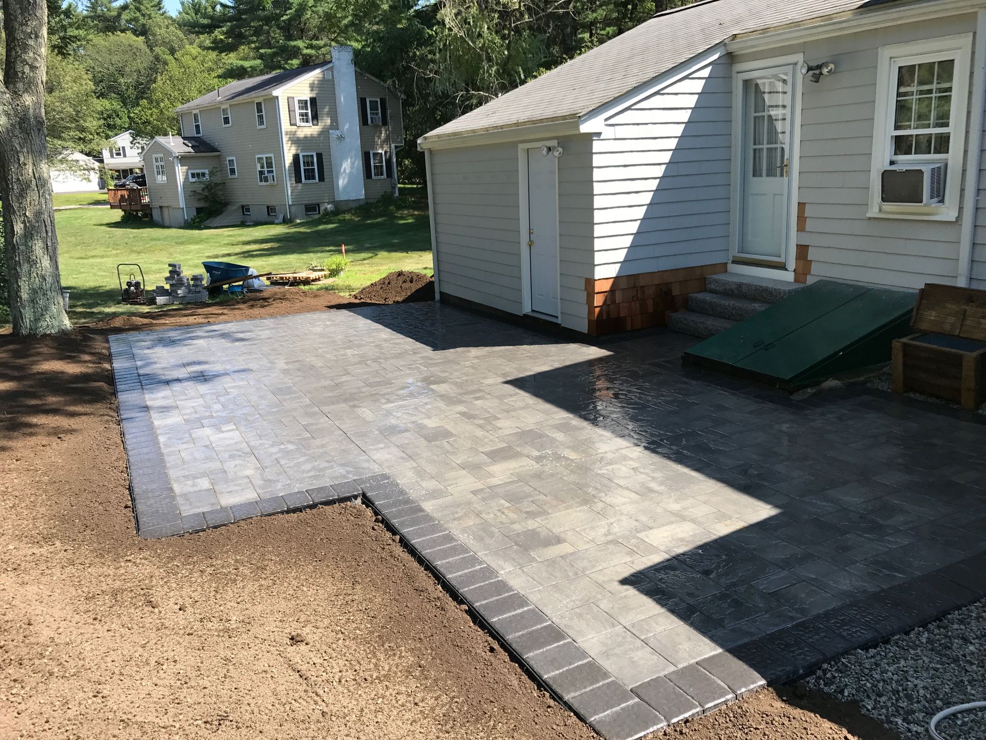A new stone paver patio beside a light gray house with a basement bulkhead, surrounded by dirt and unfinished yard.
