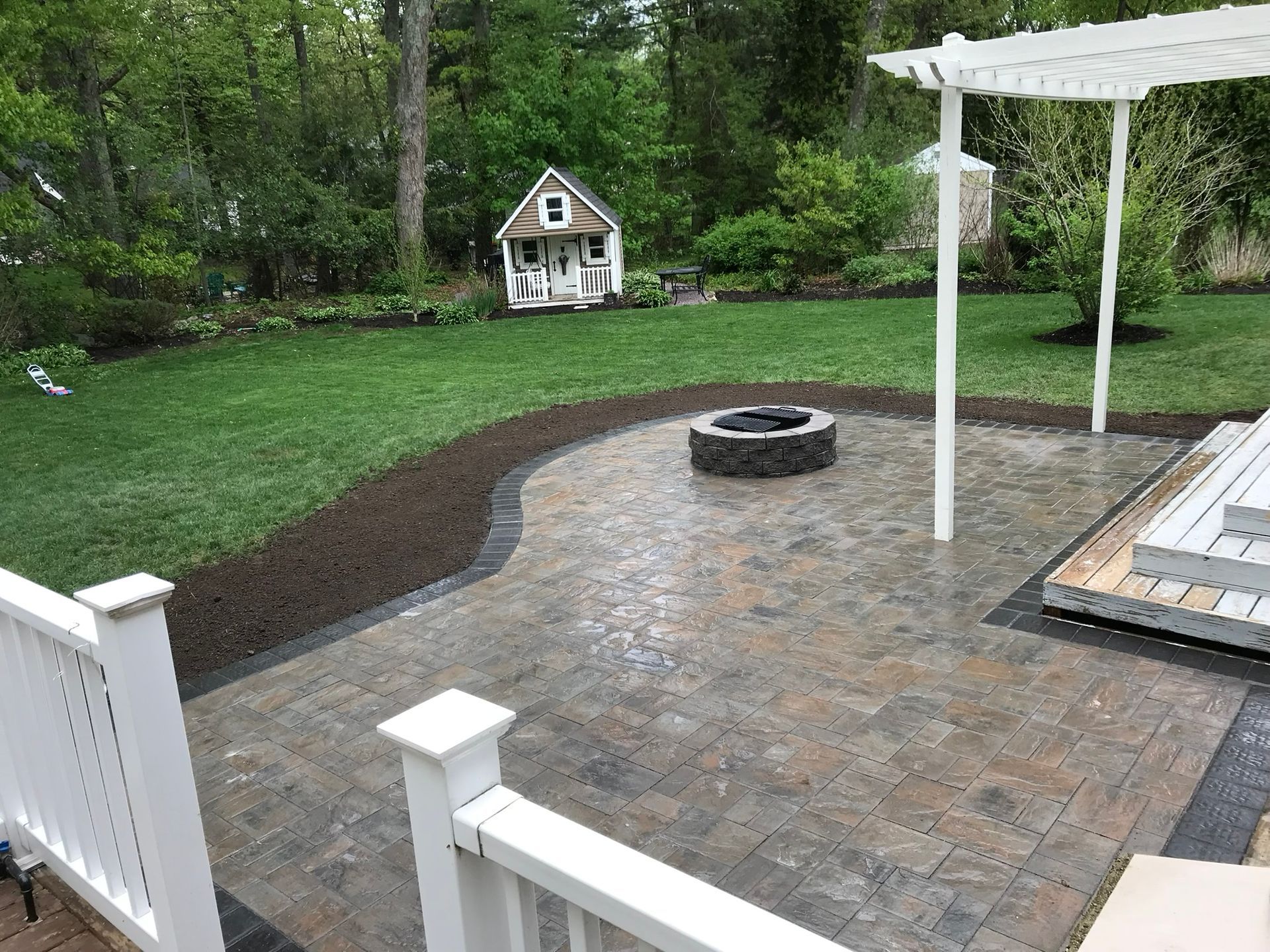 A paved stone patio with a fire pit, bordered by a lawn and a white pergola, viewed from a deck with a white railing.