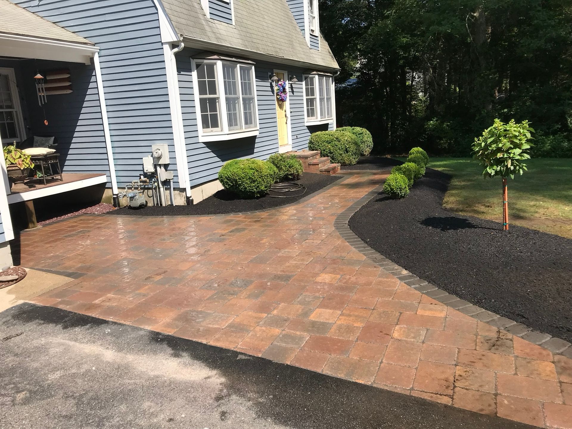 A paved brick walkway leads to the front porch of a blue house with dark mulch beds and landscaping.