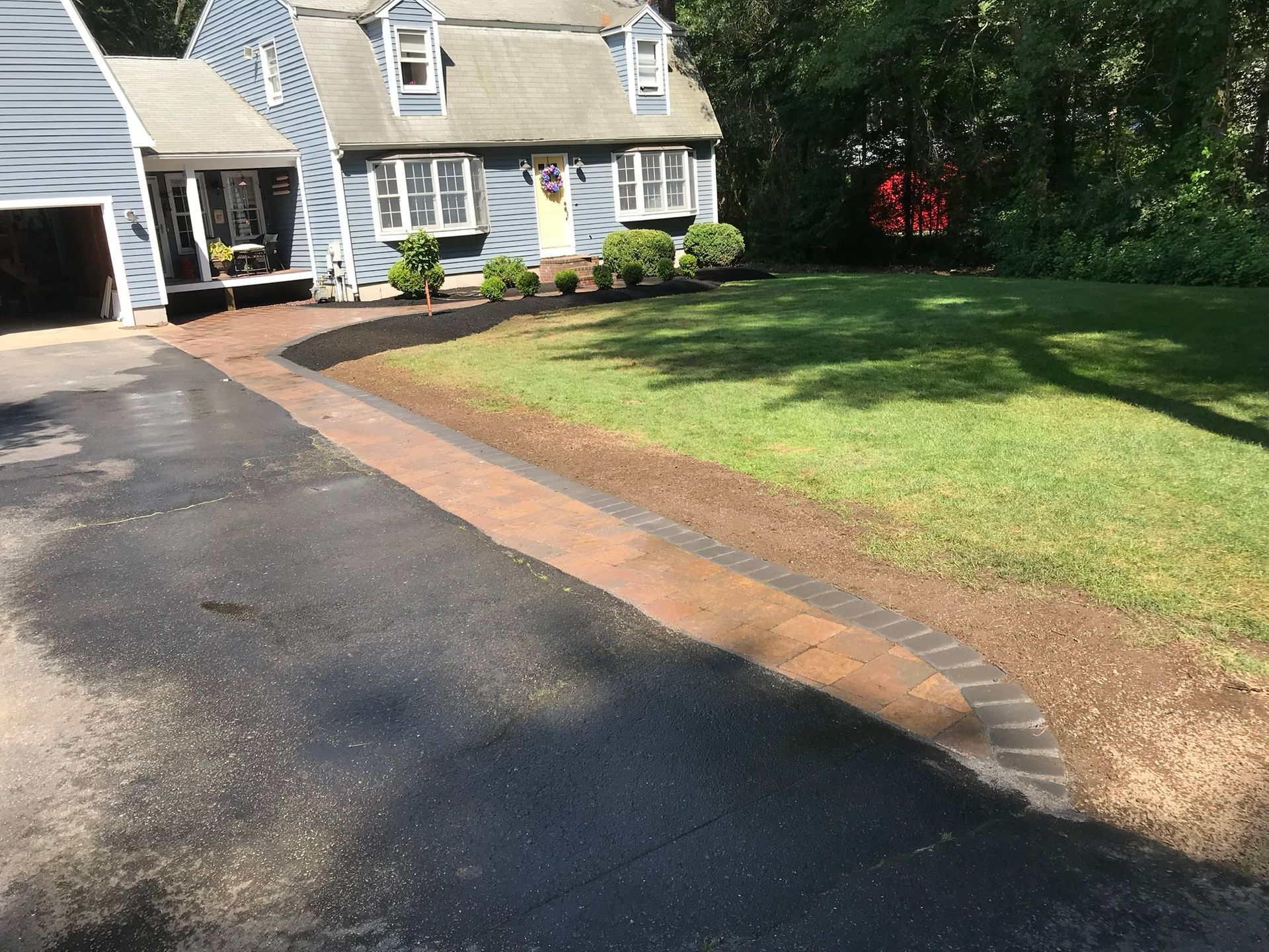 A new paver border lines an asphalt driveway in front of a blue house with a landscaped lawn and a garage.