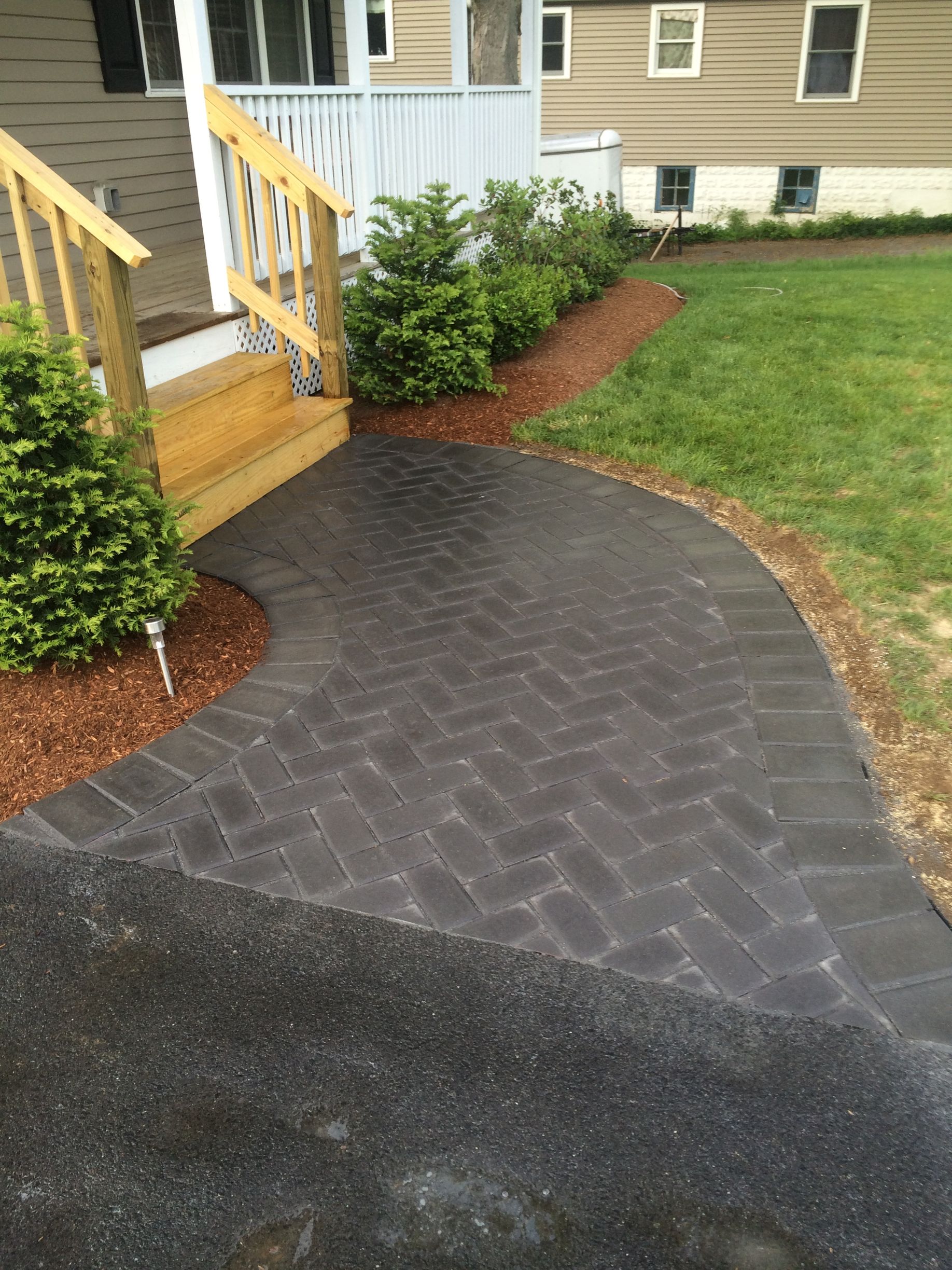 A herringbone-patterned dark gray paver walkway leads to the light wood steps of a house with green shrubs and mulch.