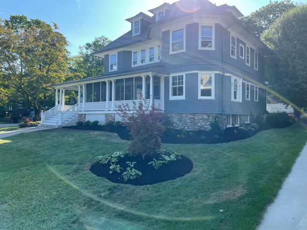 A large, grey, two-story house with a wrap-around porch, stone foundation, and a landscaped front lawn with mulch beds.