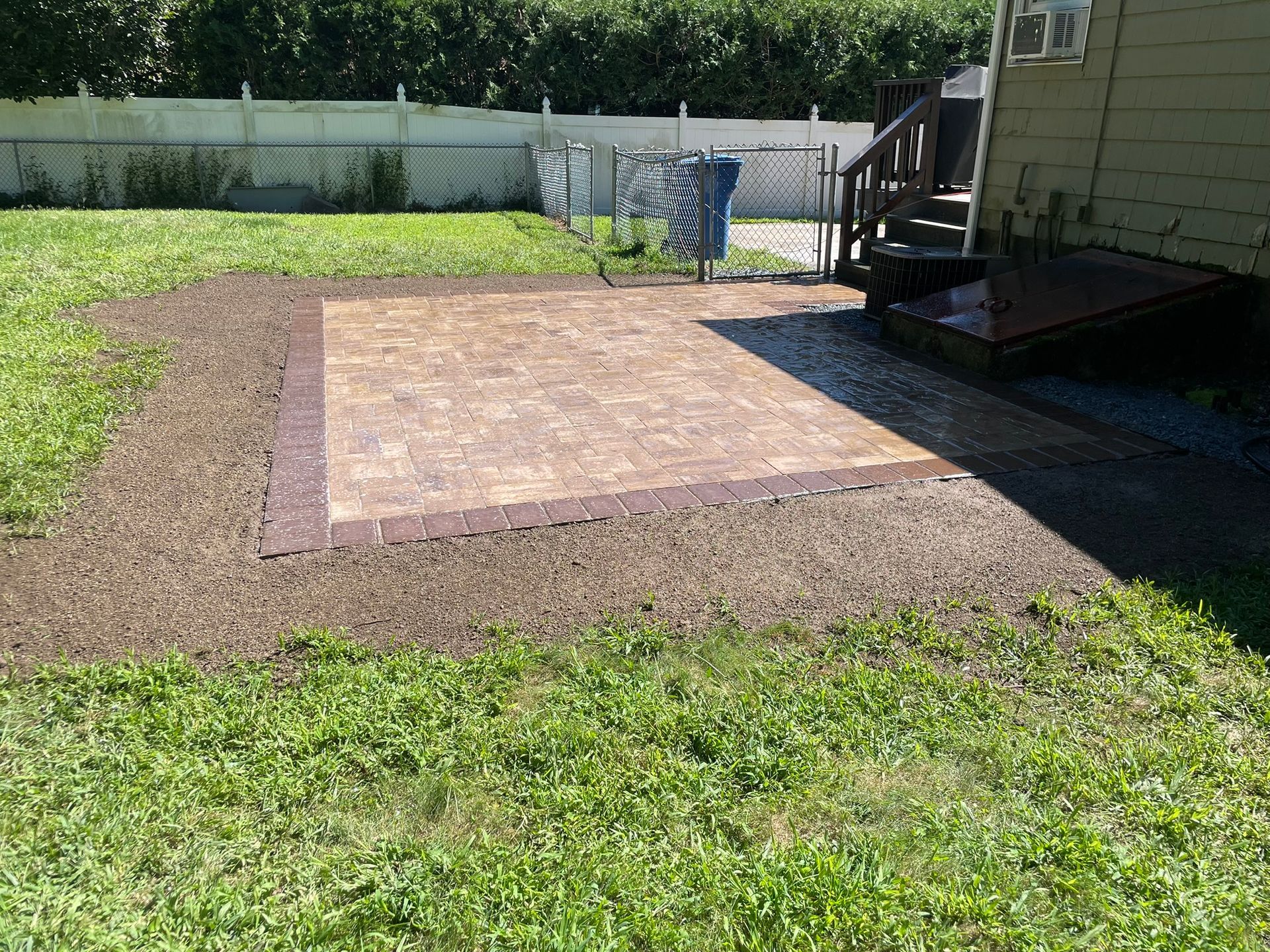 A rectangular paver patio with a dark border sits in a backyard next to a house with a cellar door and wooden steps.