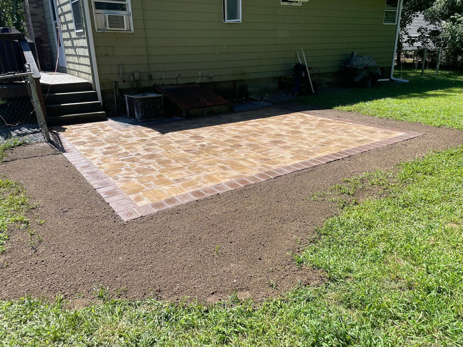 A rectangular stone paver patio with a dark border, set against the side of a house with green siding and brown mulch.