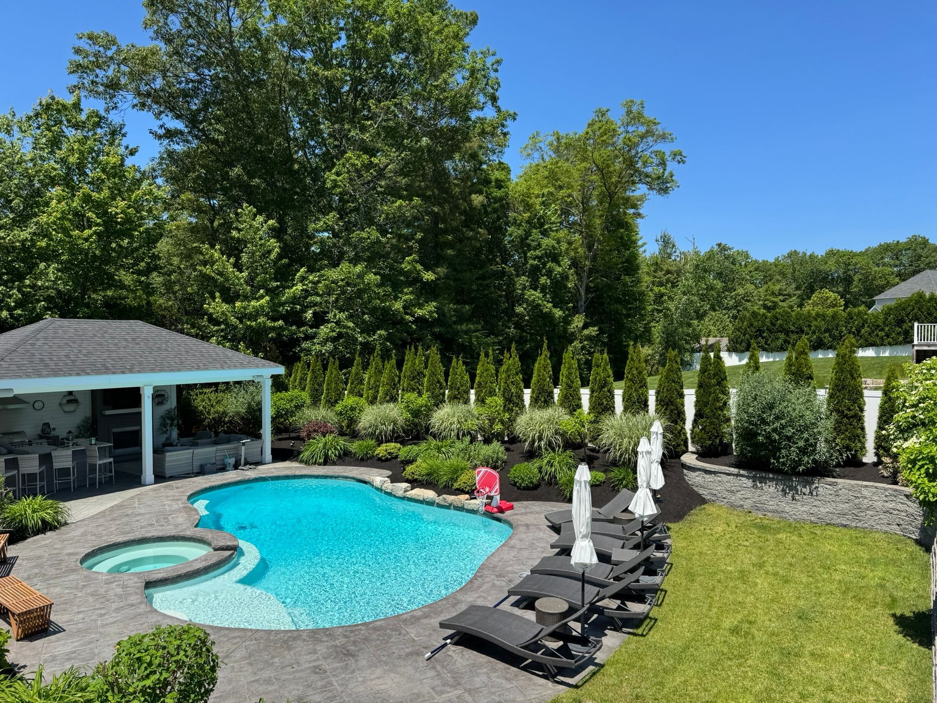 A swimming pool and hot tub next to a covered outdoor patio, surrounded by lounge chairs, landscaping, and tall trees.