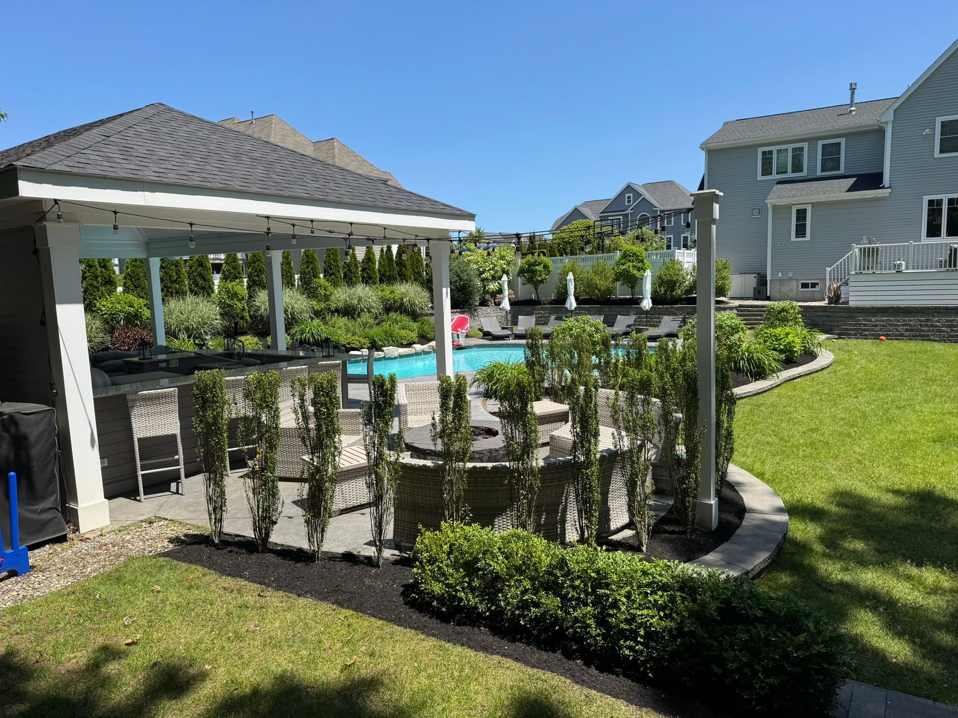 A backyard pool area featuring a shaded pavilion with seating, a stone fire pit, and landscaping on a sunny day.