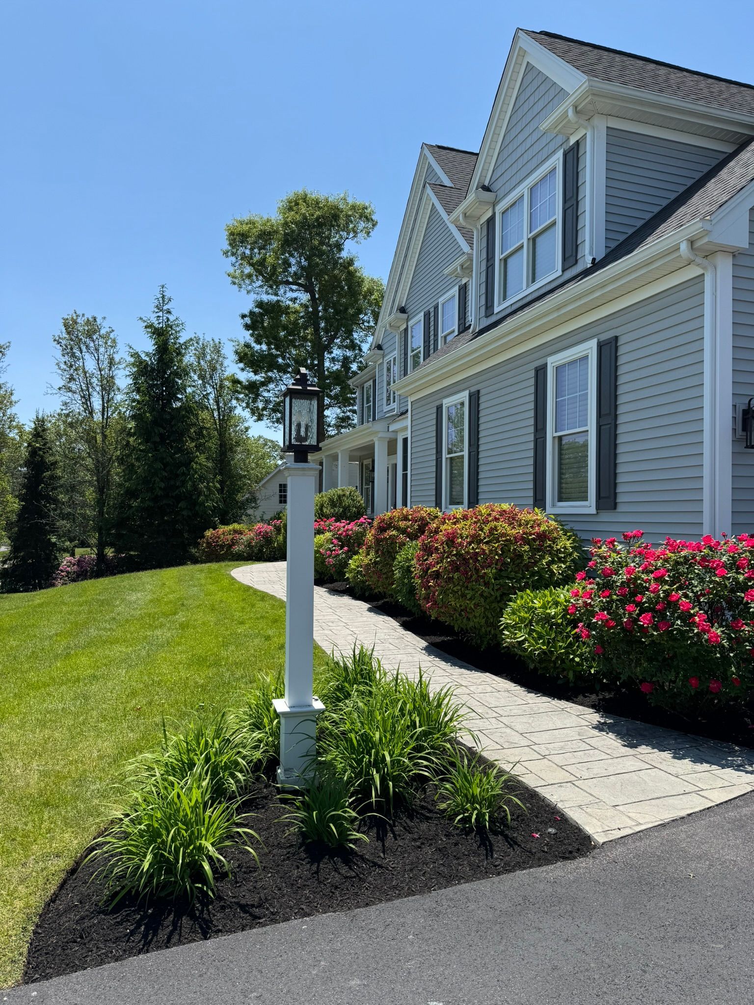 A white lamp post stands in a mulched garden bed beside a stone walkway leading to a light blue, multi-gabled house.
