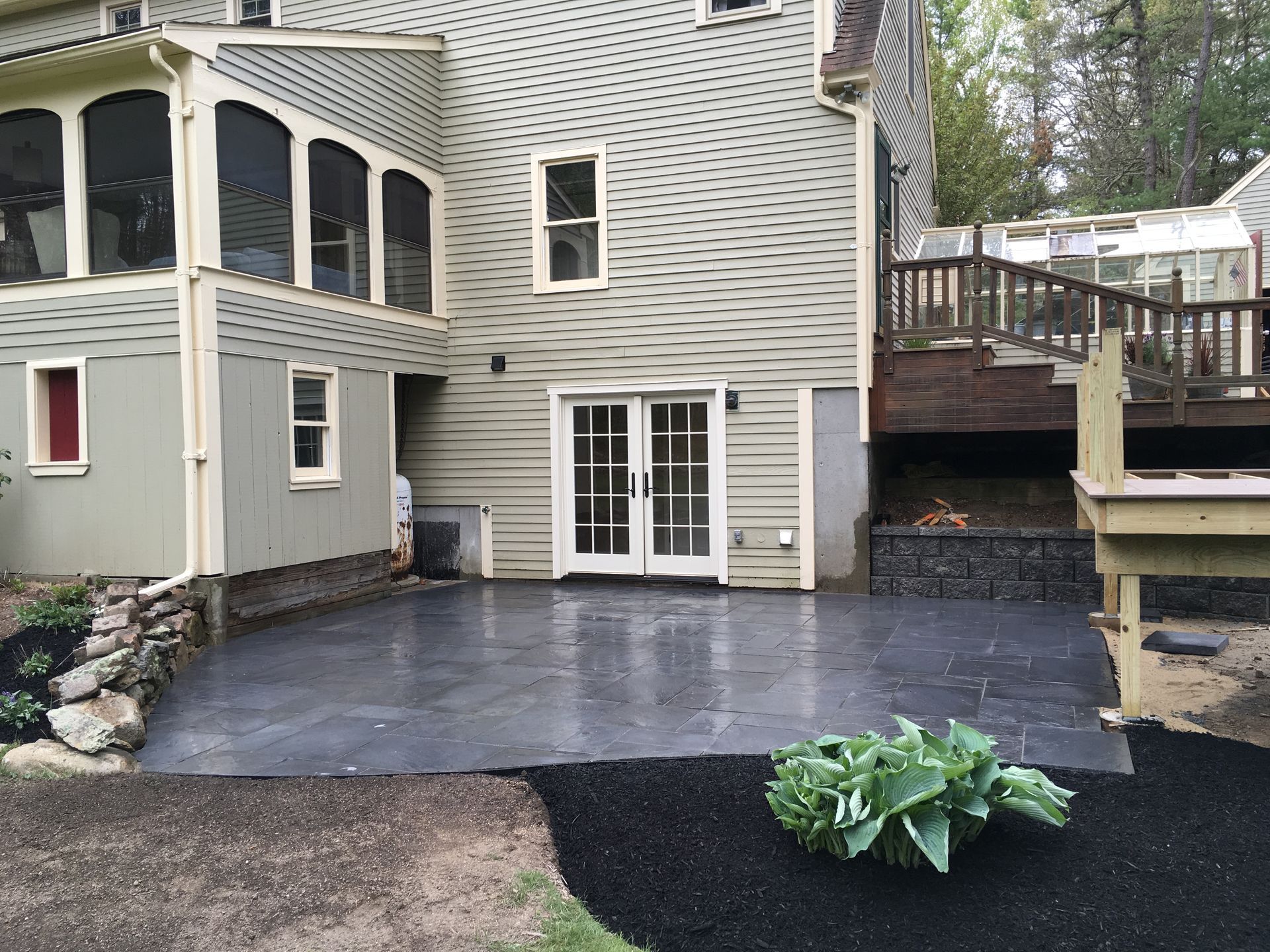 A gray stone patio outside a house with beige siding, a raised wooden deck, and a patch of mulch with a green plant.