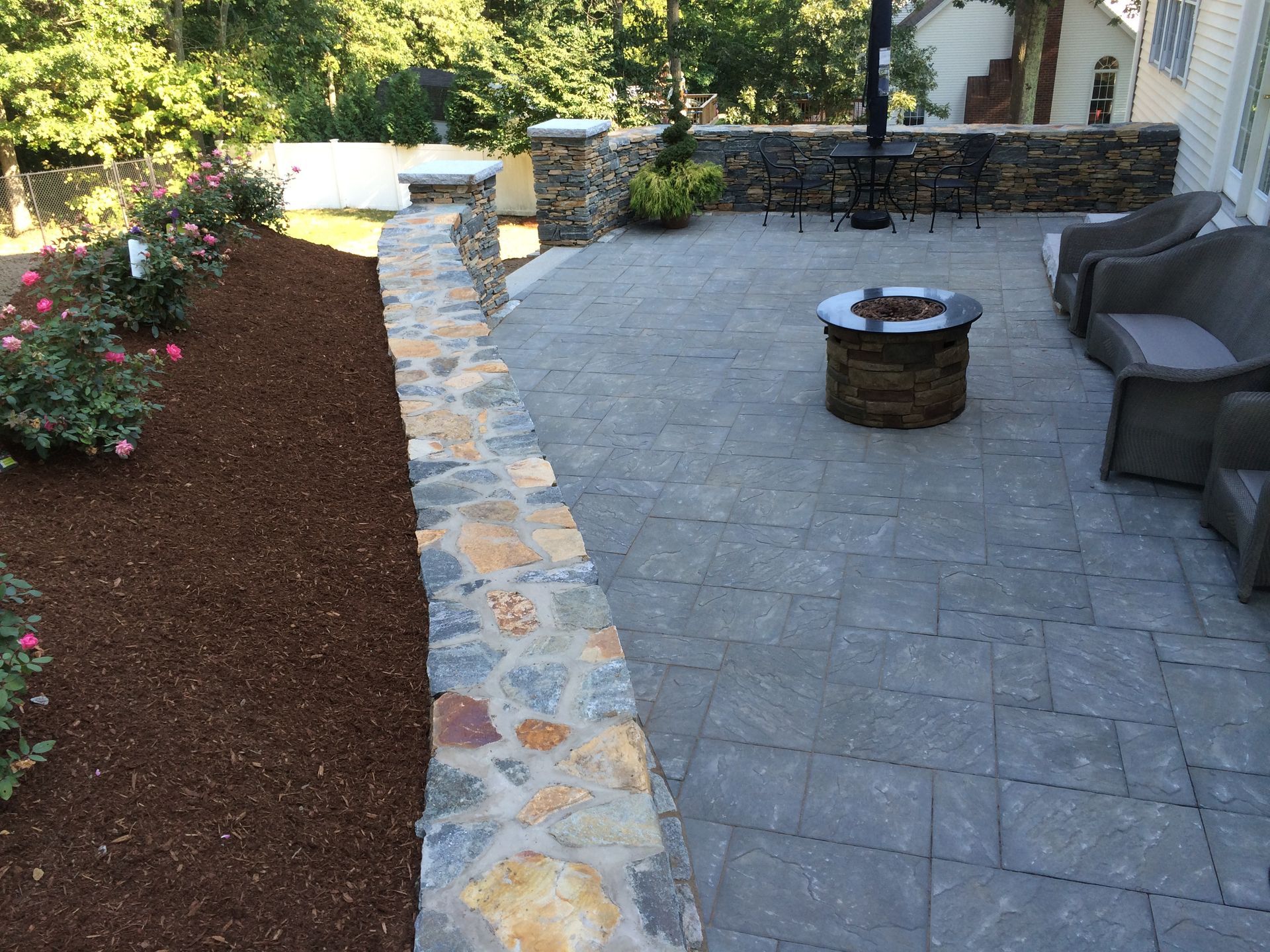 A stone patio with a fire pit, outdoor furniture, and a low stone wall next to a garden bed with roses and mulch.