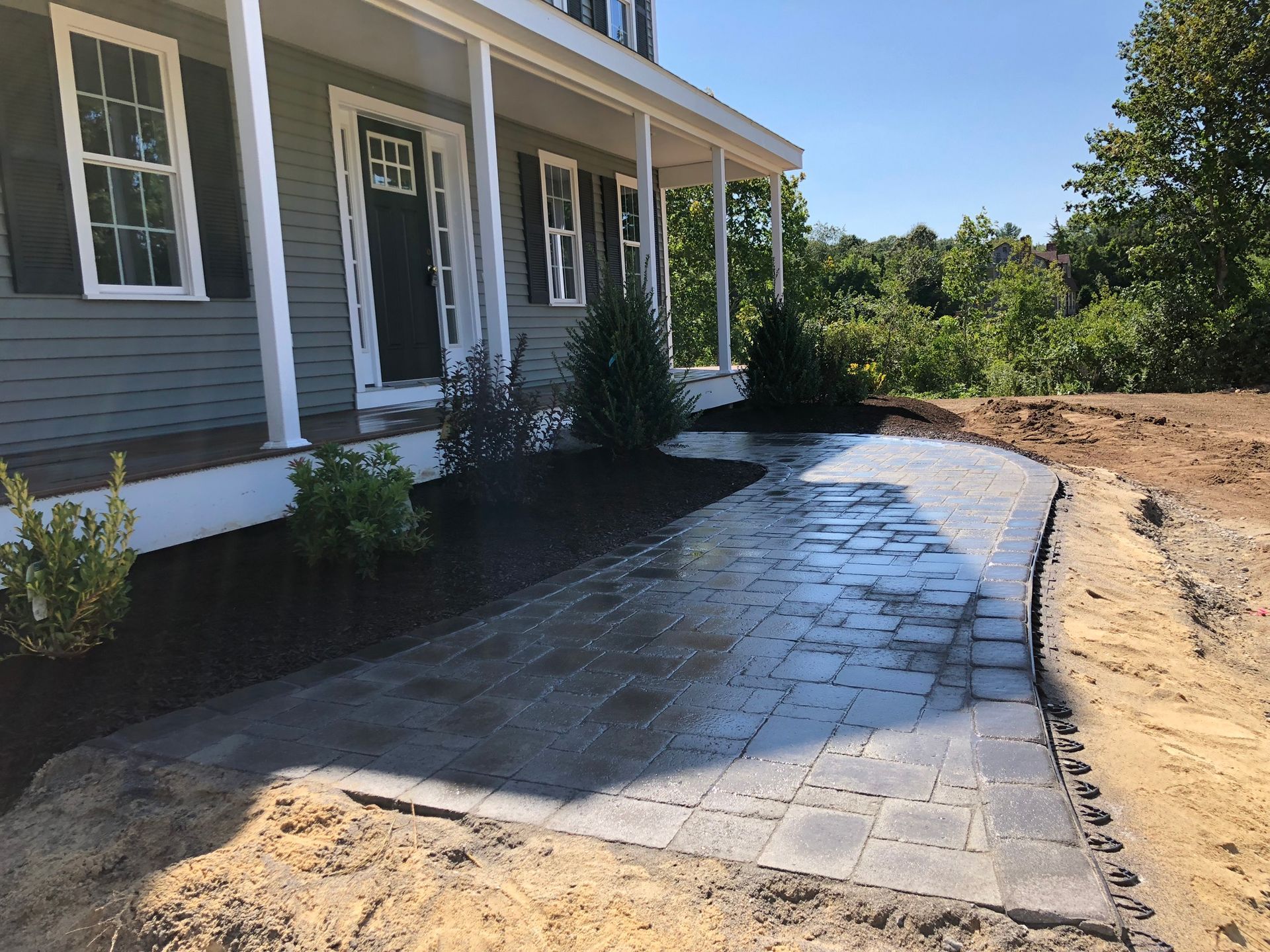 A paved stone walkway under construction leads to the front porch of a grey house with black shutters and a black door.