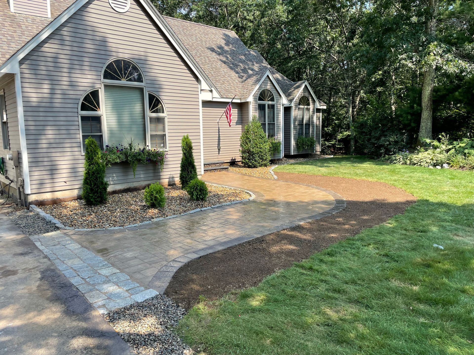 A light-gray suburban home with a newly installed paver walkway leading to the front door, bordered by mulch and greenery.