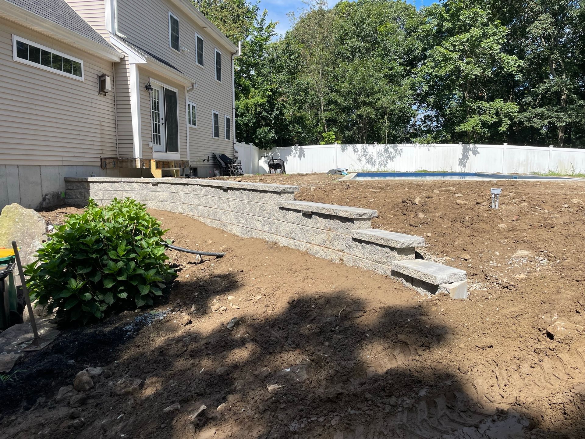 A tan house with a new stone retaining wall leading to an unfinished dirt backyard and a white fence.