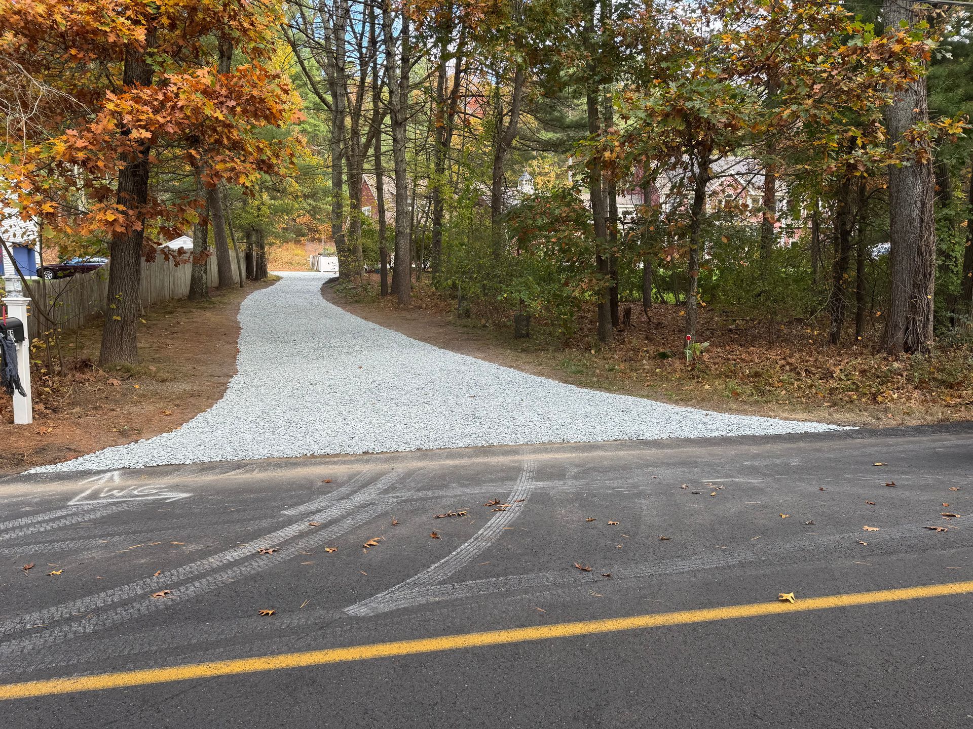 A gray gravel driveway leading from a paved road into a wooded area with fall foliage.
