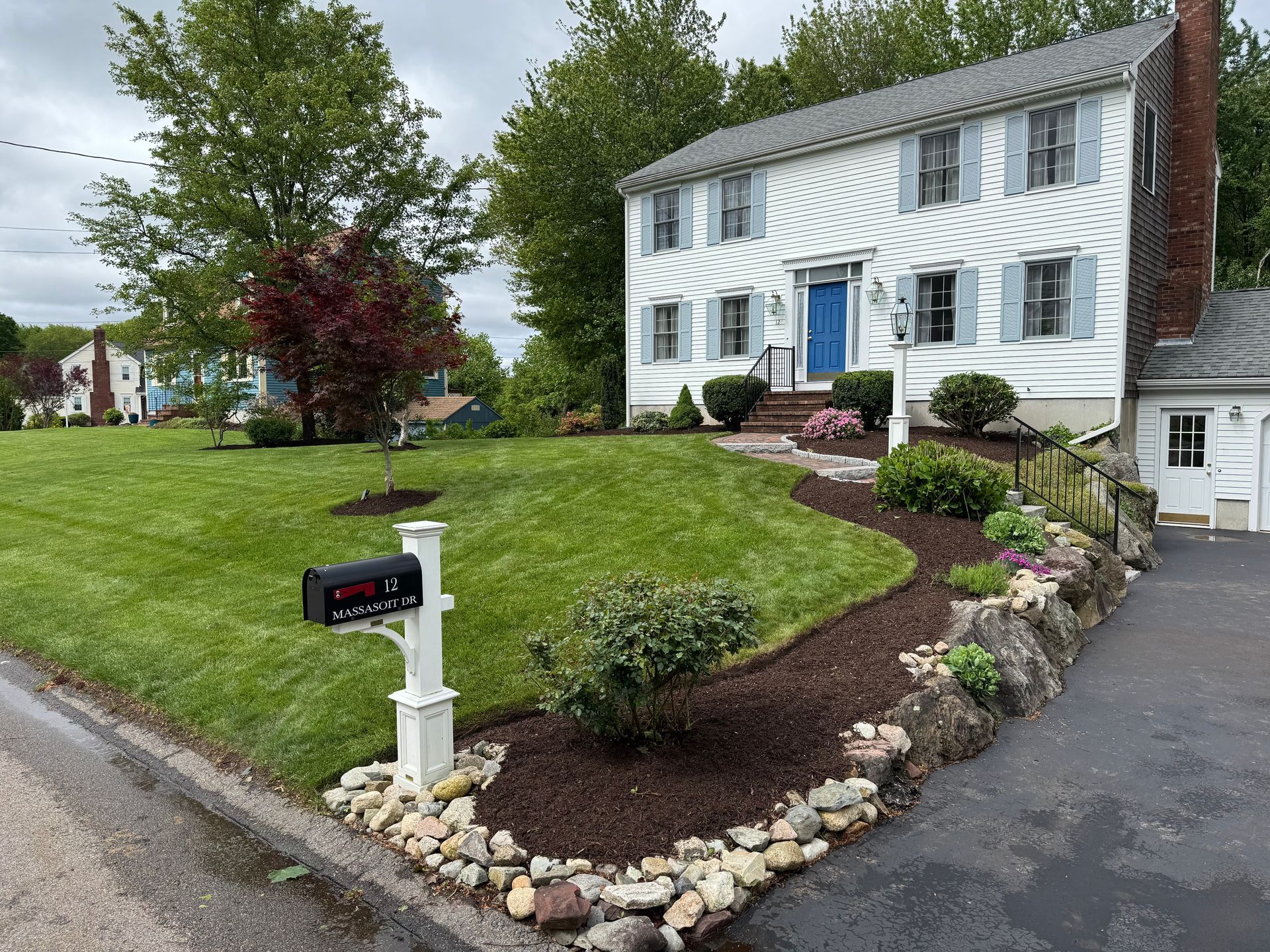 Two-story white house with blue door and shutters, lawn, landscaping, and a mailbox on the street.