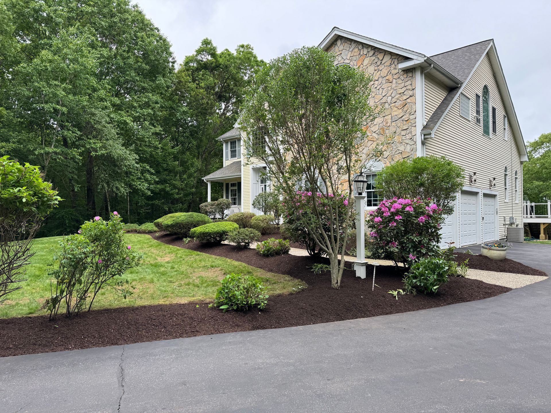 Two-story house with stone and beige siding, surrounded by trees, shrubs, and mulch-lined landscaping.