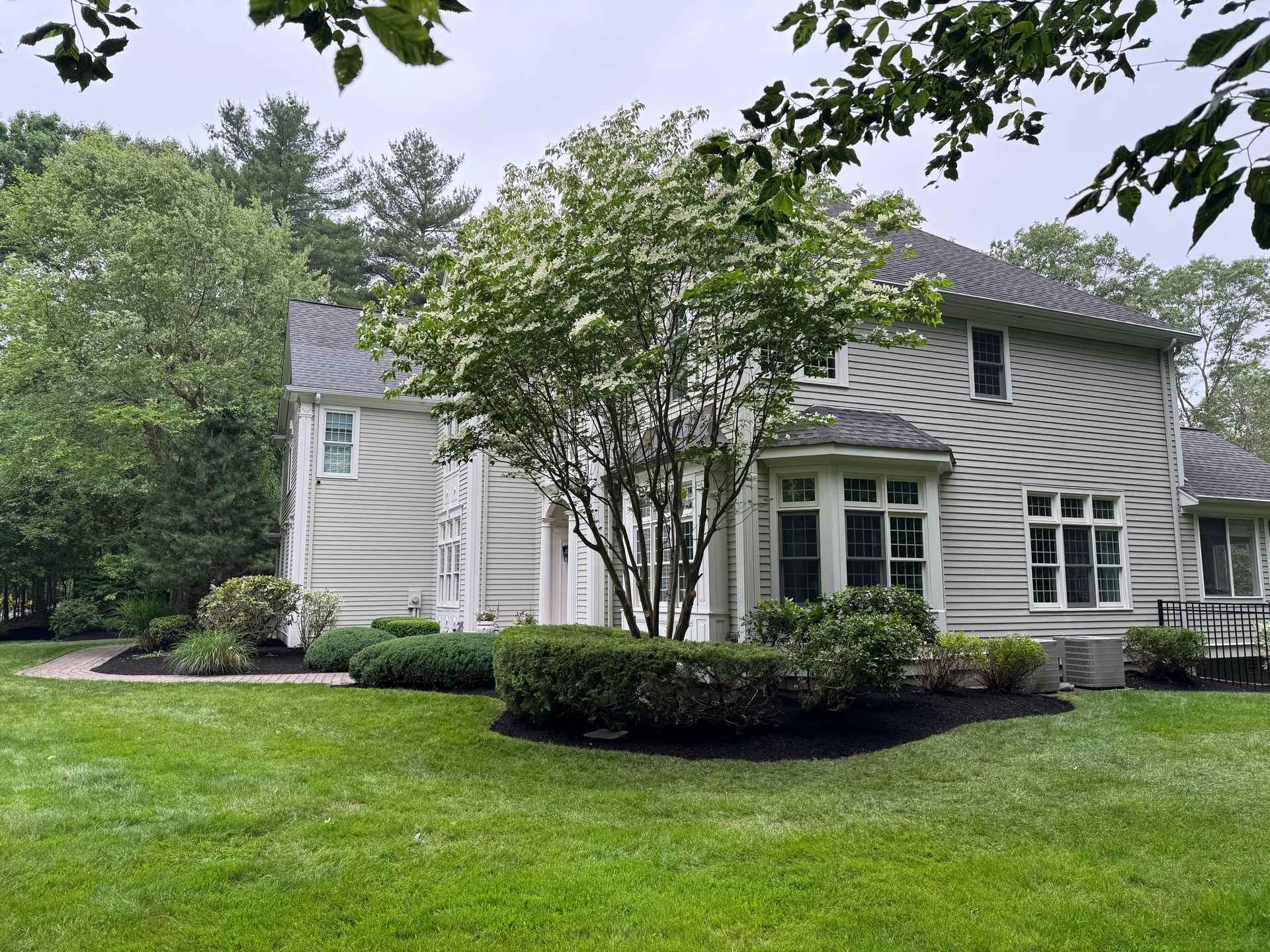 Two-story house with gray siding, surrounded by a green lawn, shrubs, and trees.