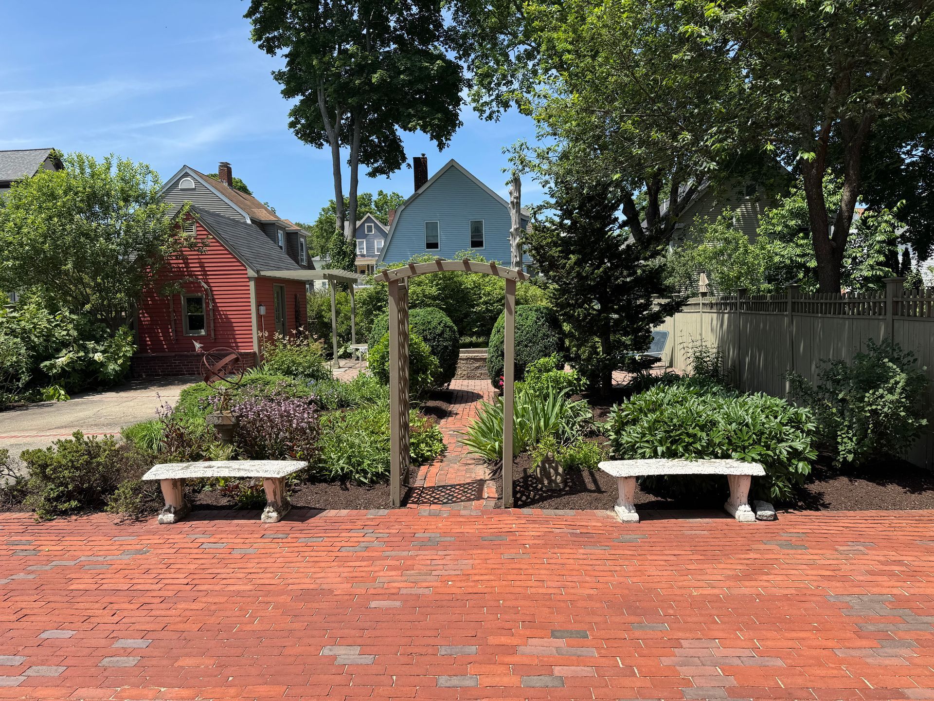 Brick pathway leads to garden with stone benches and wooden archway, with houses in the background.