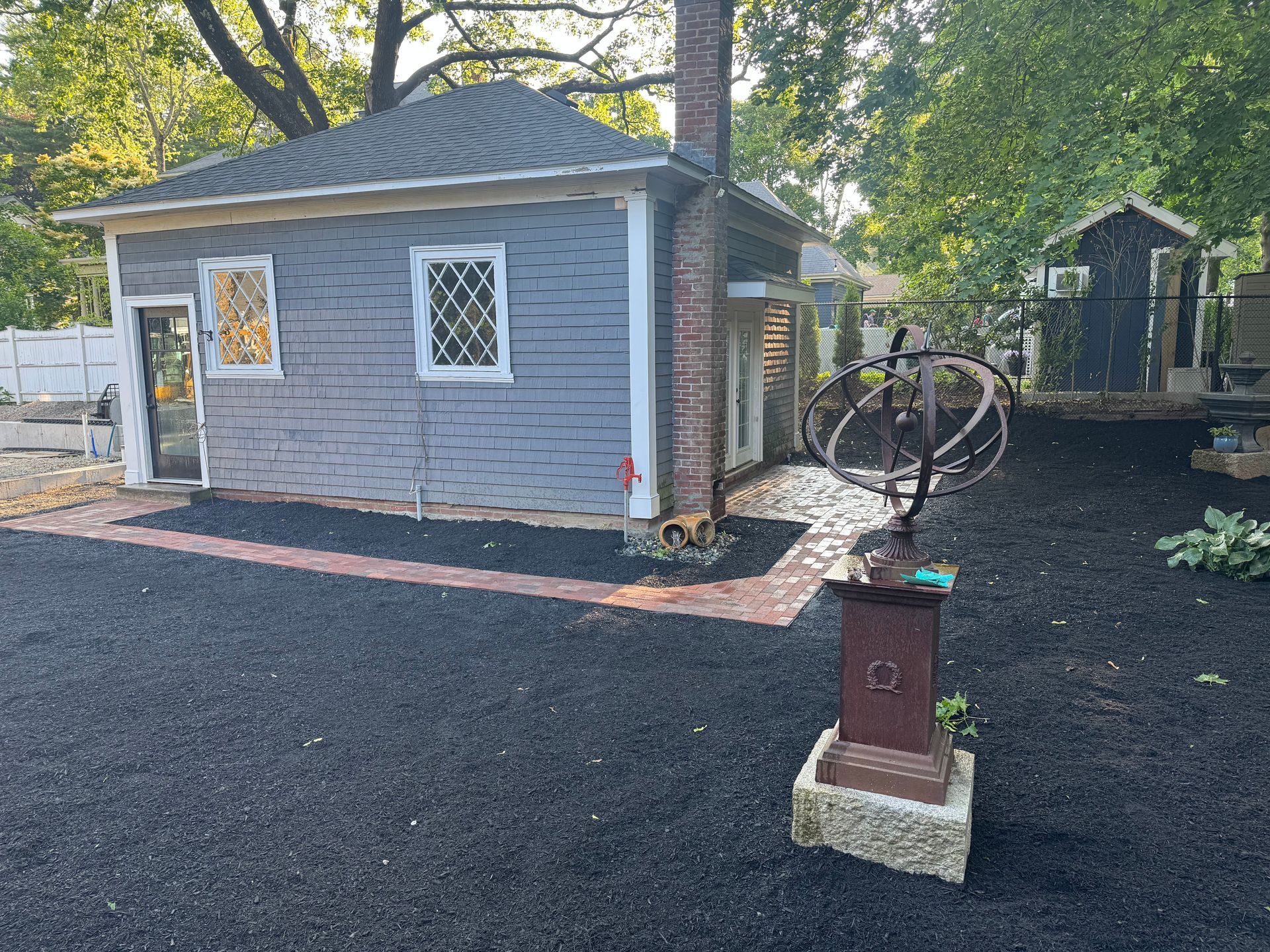A gray outbuilding with a brick chimney and trellis windows, set in a yard with a sundial and freshly laid dark mulch.