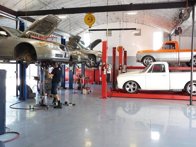 Mechanic working under a silver car raised on a lift in a garage. Two other cars on lifts are also visible.