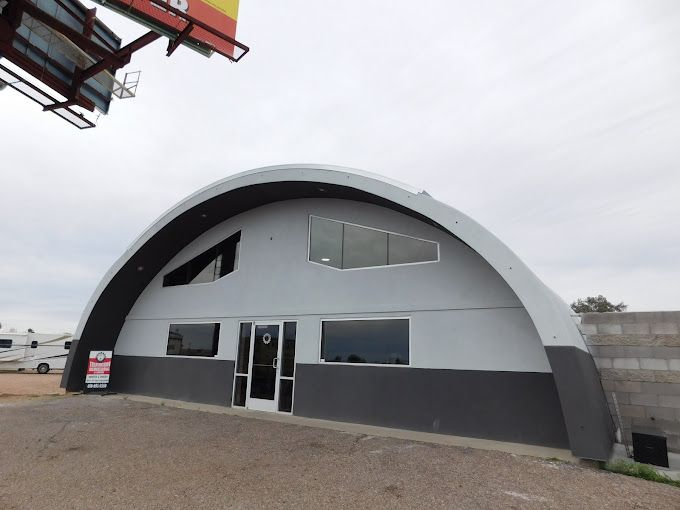 Quonset hut-style building, gray exterior with dark gray base, large arched roof. A for sale sign is visible.