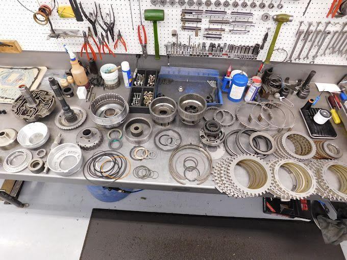 Disassembled car transmission parts neatly arranged on a metal workbench in a workshop, surrounded by tools.