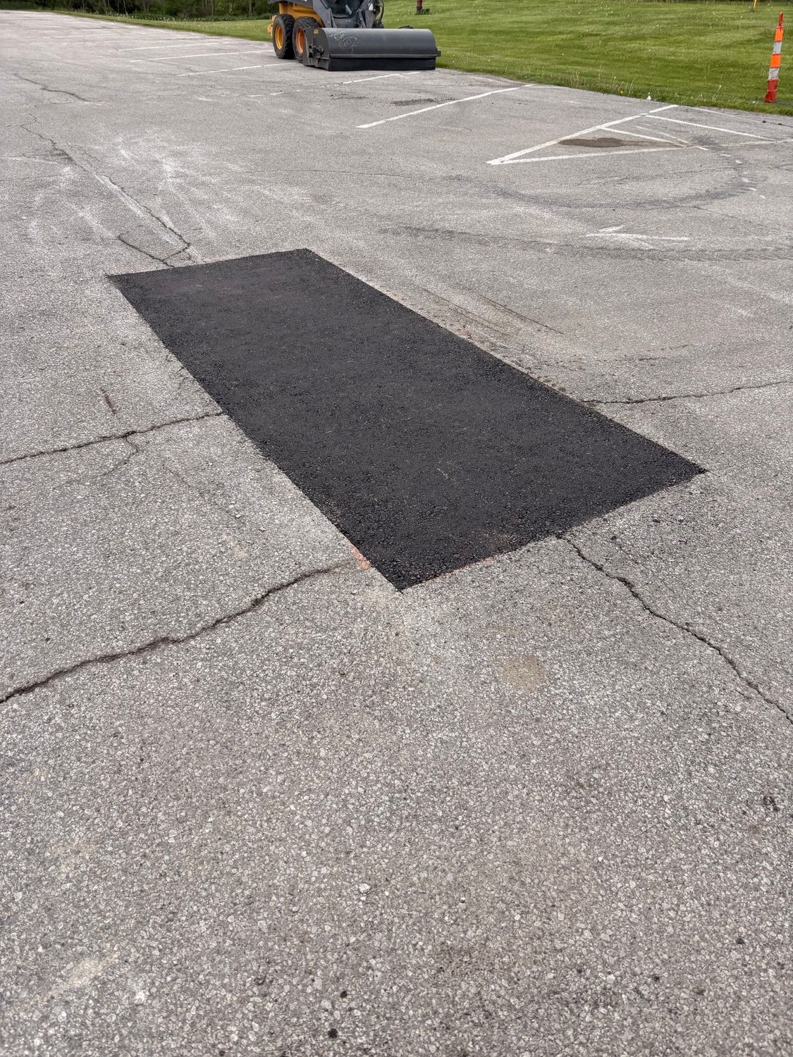 Fresh black asphalt patch on cracked gray pavement near a road roller in a parking lot
