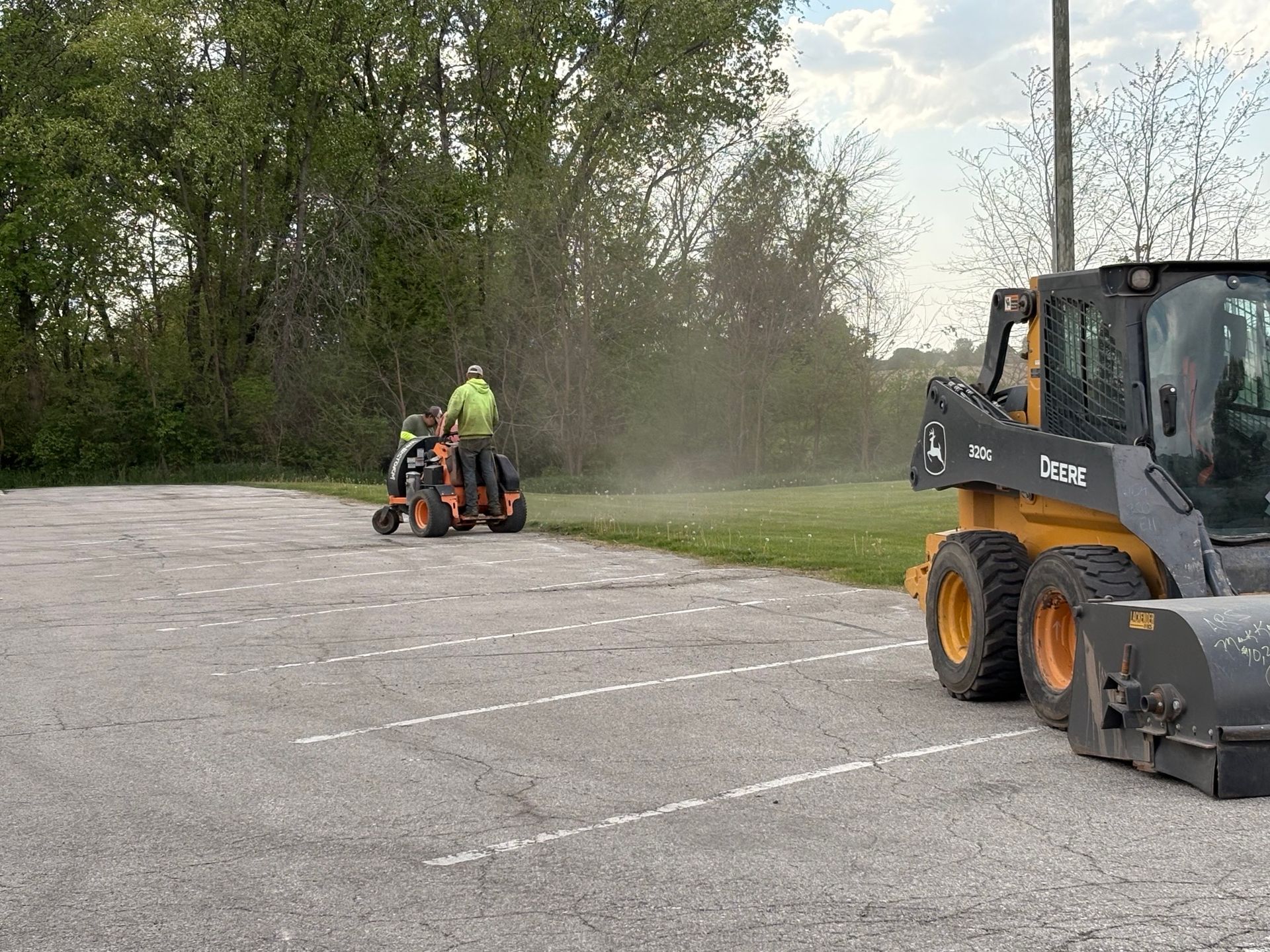 Worker operating an orange mower on a dusty paved lot near a black skid steer.