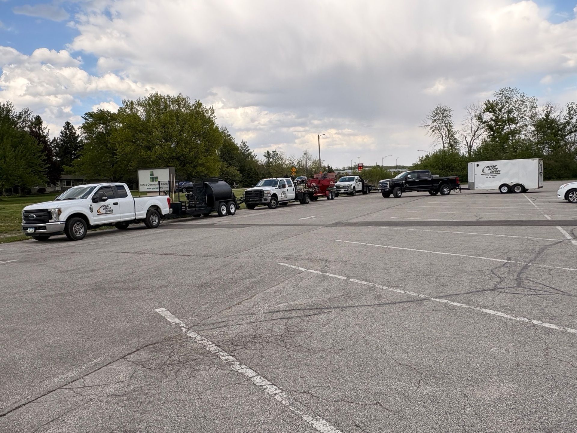 Parking lot with several parked vehicles under a cloudy sky, including white trucks and trailers.