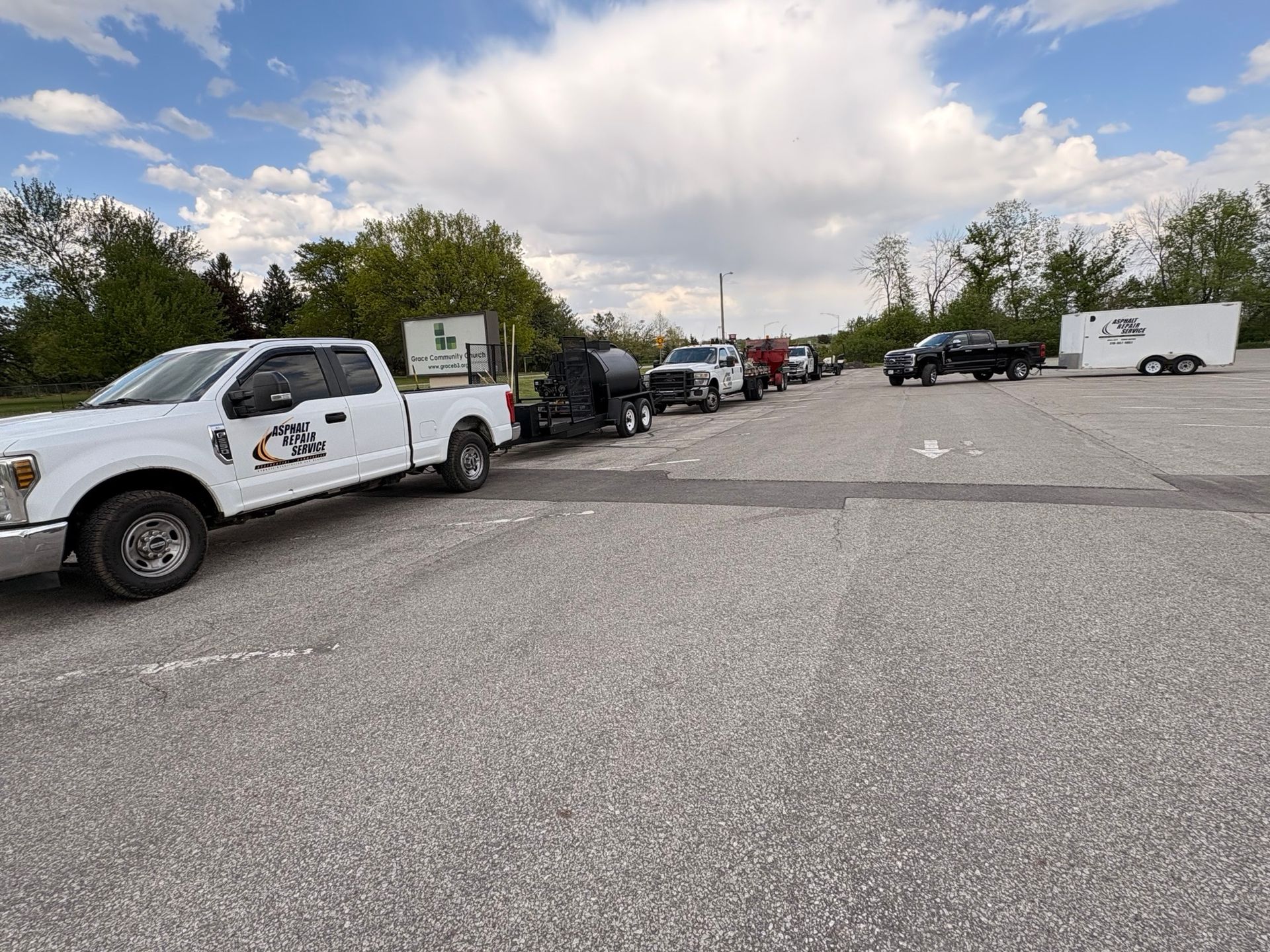 Parked work trucks and trailers along a wide gravel lot under a partly cloudy sky