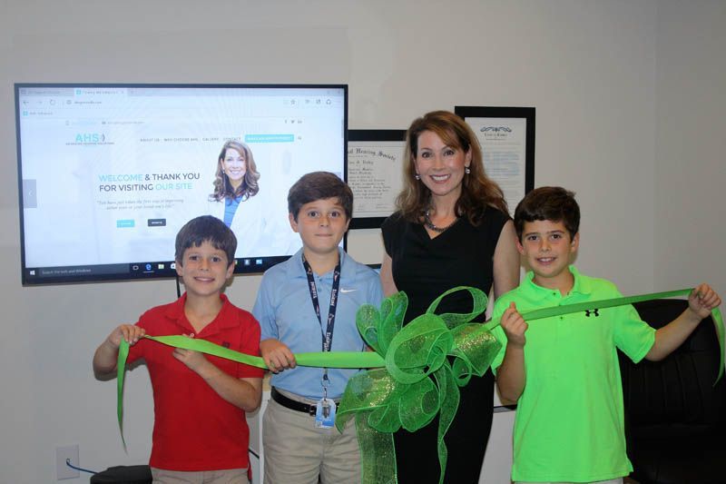 Woman and three boys cutting a green ribbon in front of a computer screen; indoors.