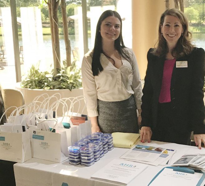 Two women at a table with promotional materials. One wears a white blouse, the other a black blazer. Indoor setting.