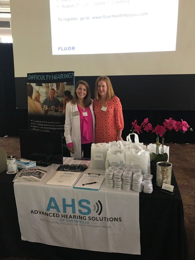 Two women at a table for Advanced Hearing Solutions; display of products, pink shirts, black tablecloth.