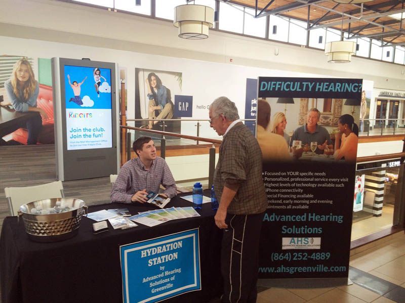 Man talking to a person at a hearing solutions booth in a mall. 