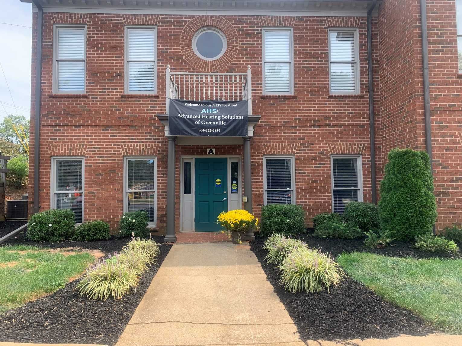 Two-story brick building with turquoise door, balcony sign, and flower beds.