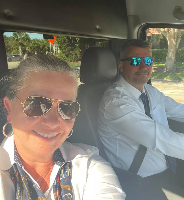 Two people wearing sunglasses in a car, smiling at the camera with a sunny street outside the window