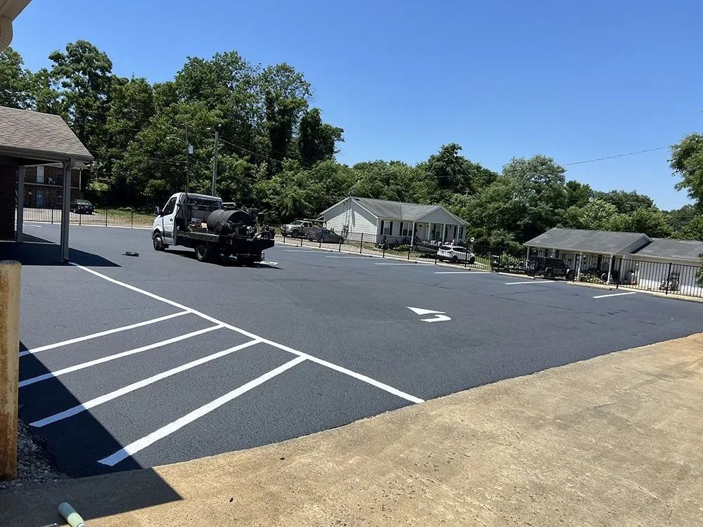 Newly paved asphalt parking lot with white painted parking space lines and a truck. Houses and trees are in the background.