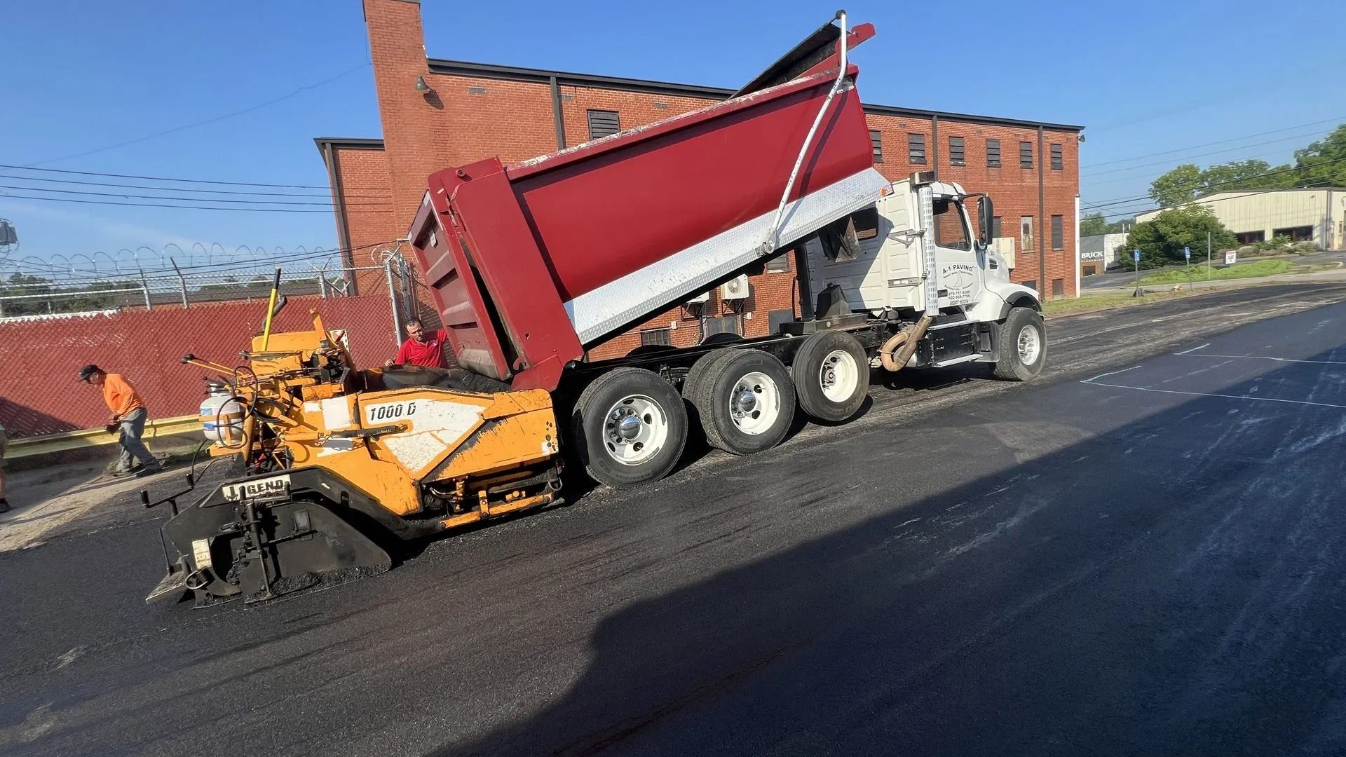 A dump truck unloading asphalt into a paving machine on a street. The scene is in front of a red brick building.