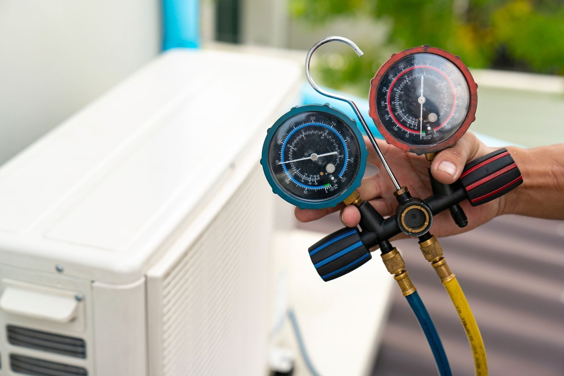 Hand holding HVAC manifold gauges with blue and red dials beside an outdoor air conditioner unit