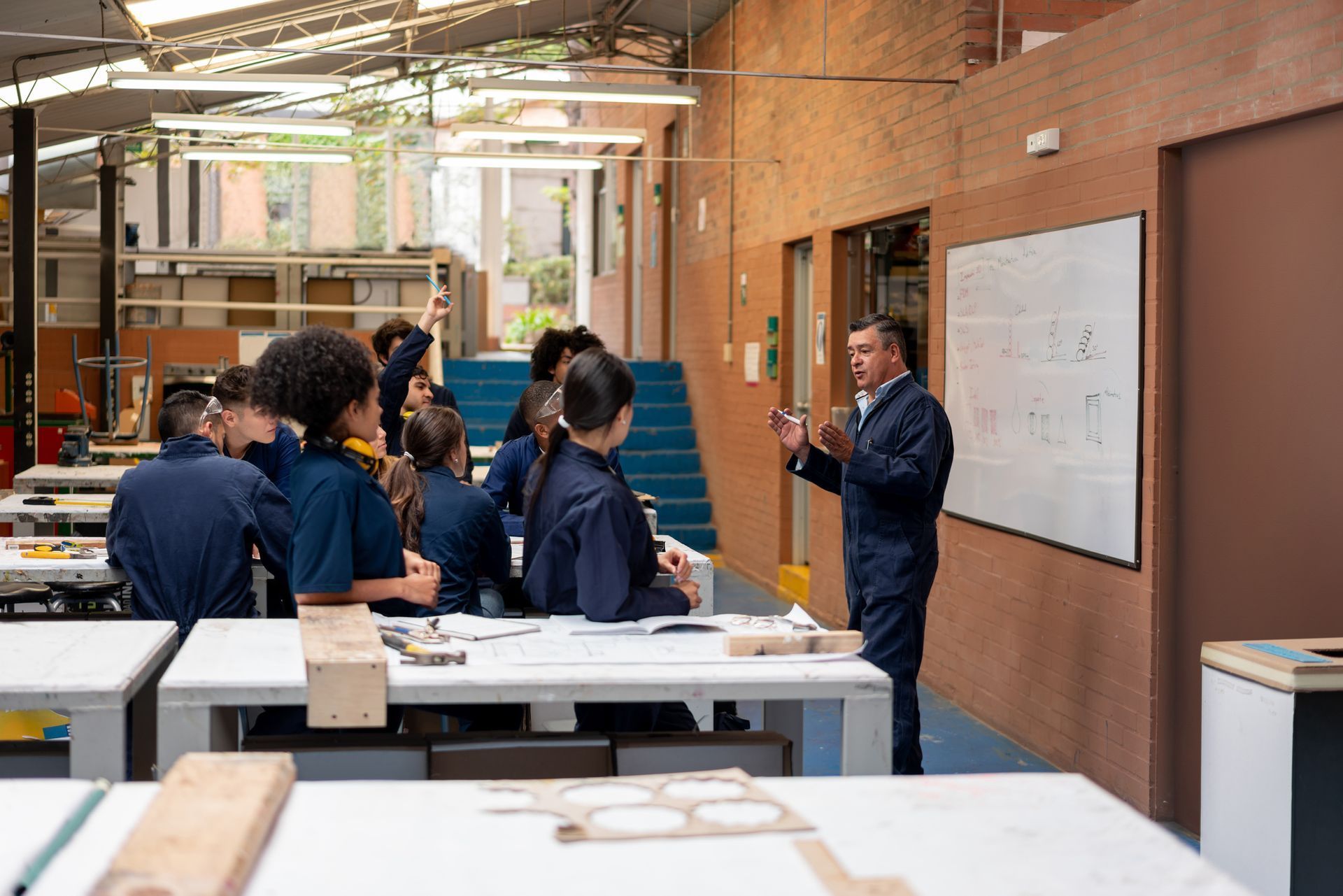 Teacher speaking to students in a bright workshop classroom with tables and wall charts.