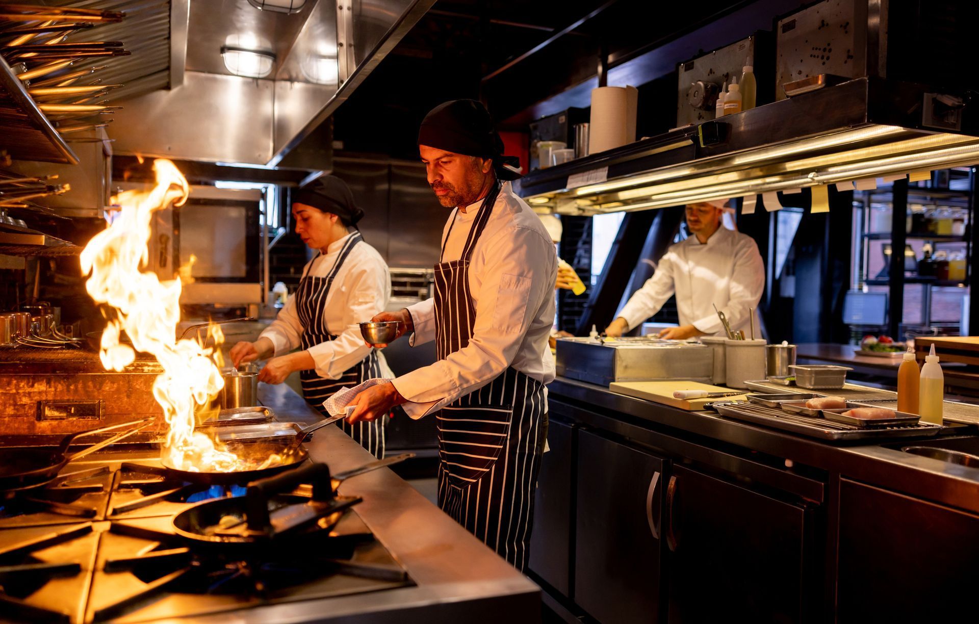 Chefs cooking in a restaurant kitchen, with a large flame flaring from a stovetop pan