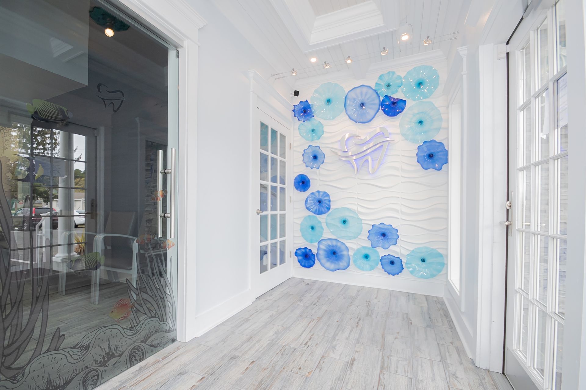 White hallway with pale wood floors, a decorative blue wall sculpture, and glass doors.