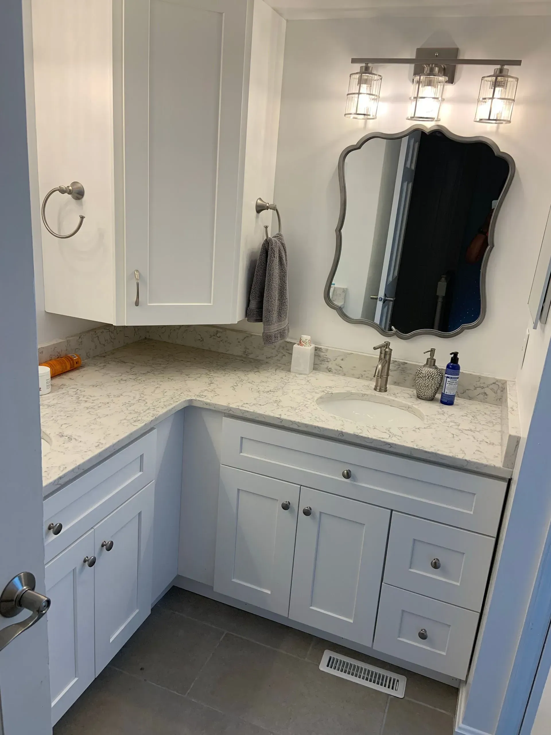 White bathroom with angled countertop, vanity, mirror, and cabinet. Silver hardware and lighting.